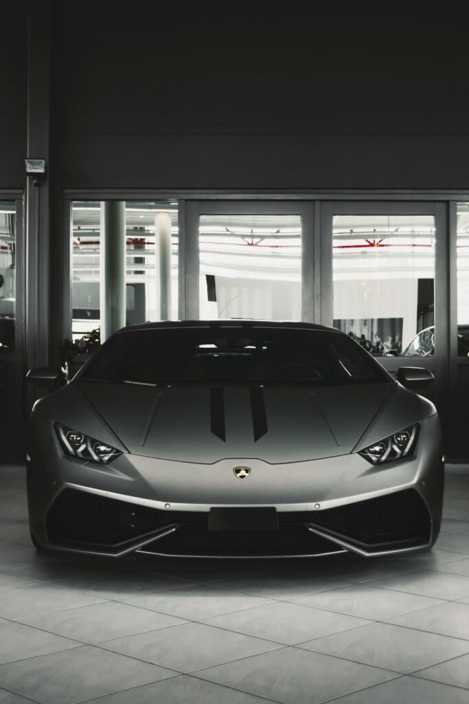 Front view of a sleek, dark grey Lamborghini Huracán parked indoors on a tiled floor, with two subtle black racing stripes down the center of the hood. The car is positioned in front of a glass wall.