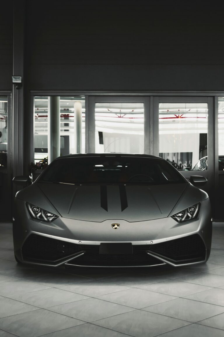 Front view of a sleek, dark grey Lamborghini Huracán parked indoors on a tiled floor, with two subtle black racing stripes down the center of the hood. The car is positioned in front of a glass wall.