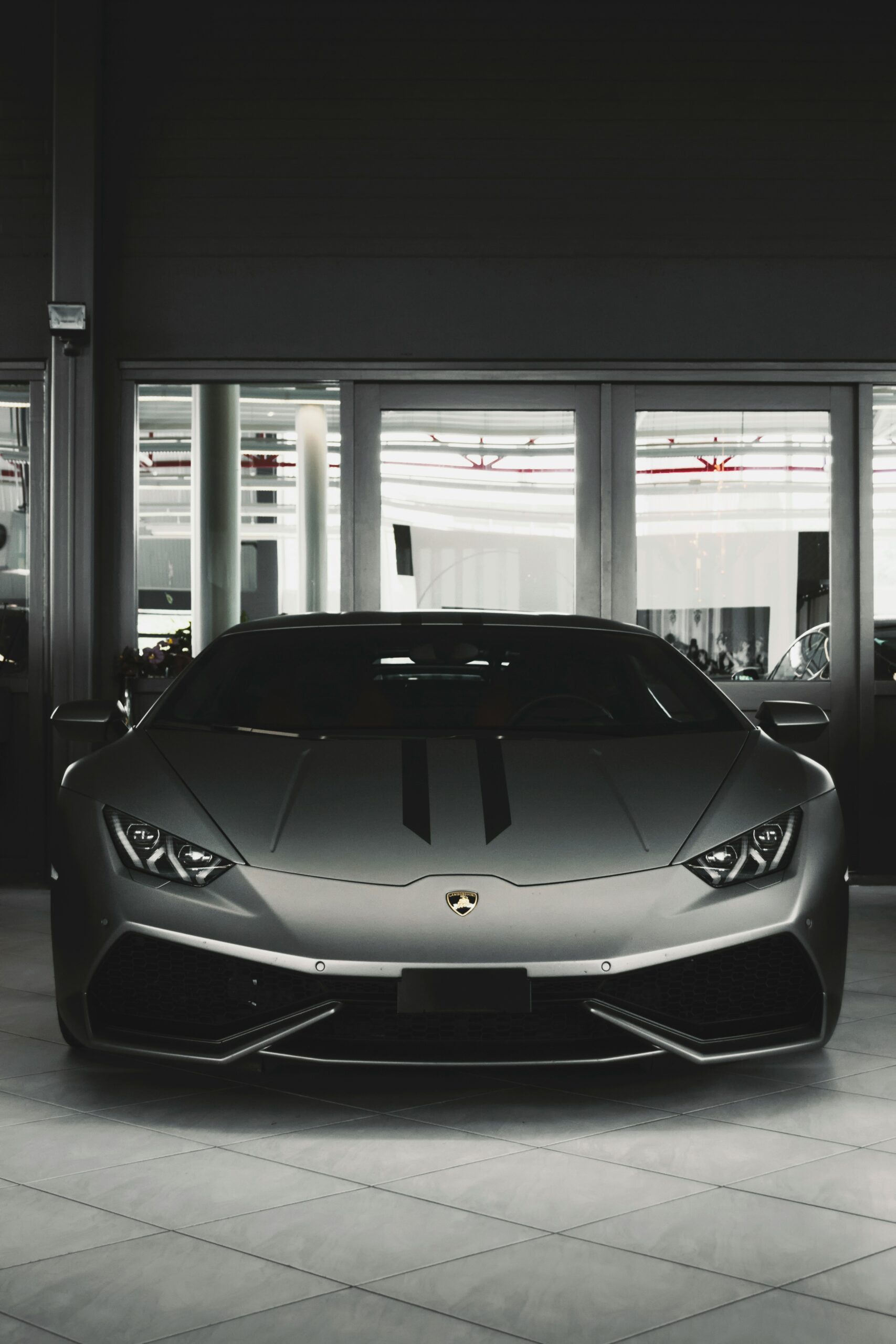Front view of a sleek, dark grey Lamborghini Huracán parked indoors on a tiled floor, with two subtle black racing stripes down the center of the hood. The car is positioned in front of a glass wall.