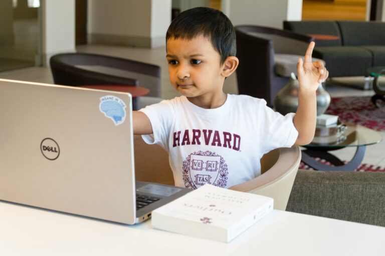 A young boy in a Harvard t-shirt sits at a laptop with a book, raising his hand in what looks like a virtual class.