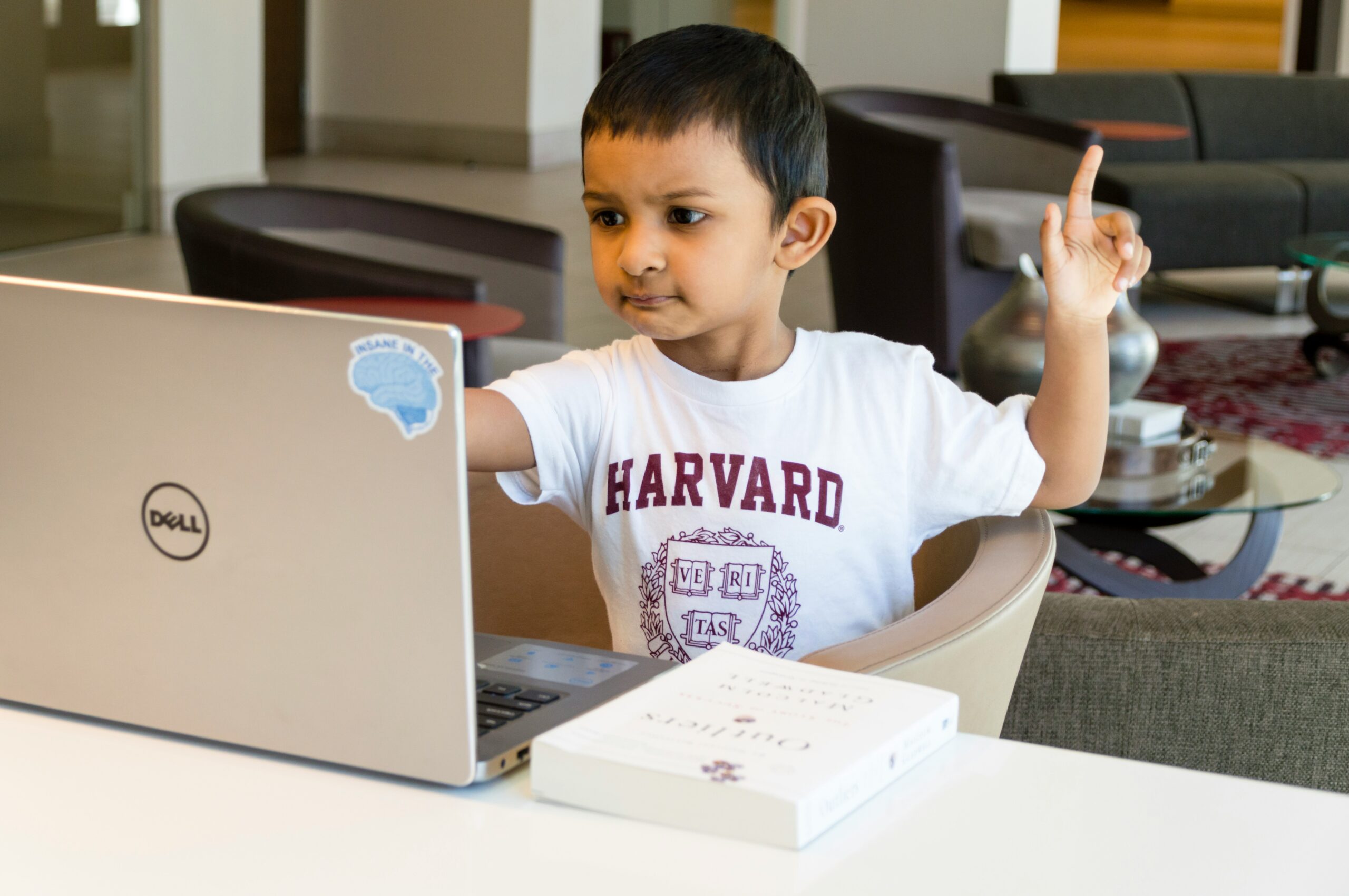 A young boy in a Harvard t-shirt sits at a laptop with a book, raising his hand in what looks like a virtual class.