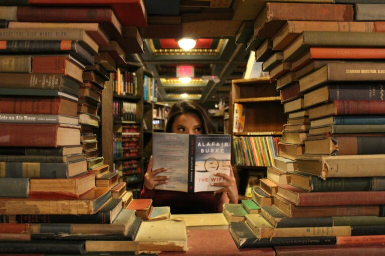 A woman is reading IDP full form partially obscured by an open book she is holding, peeking over the top. She is framed by large, uneven stacks of books on both sides, creating a tunnel-like view into a warmly lit, slightly blurred background of a library or bookstore.