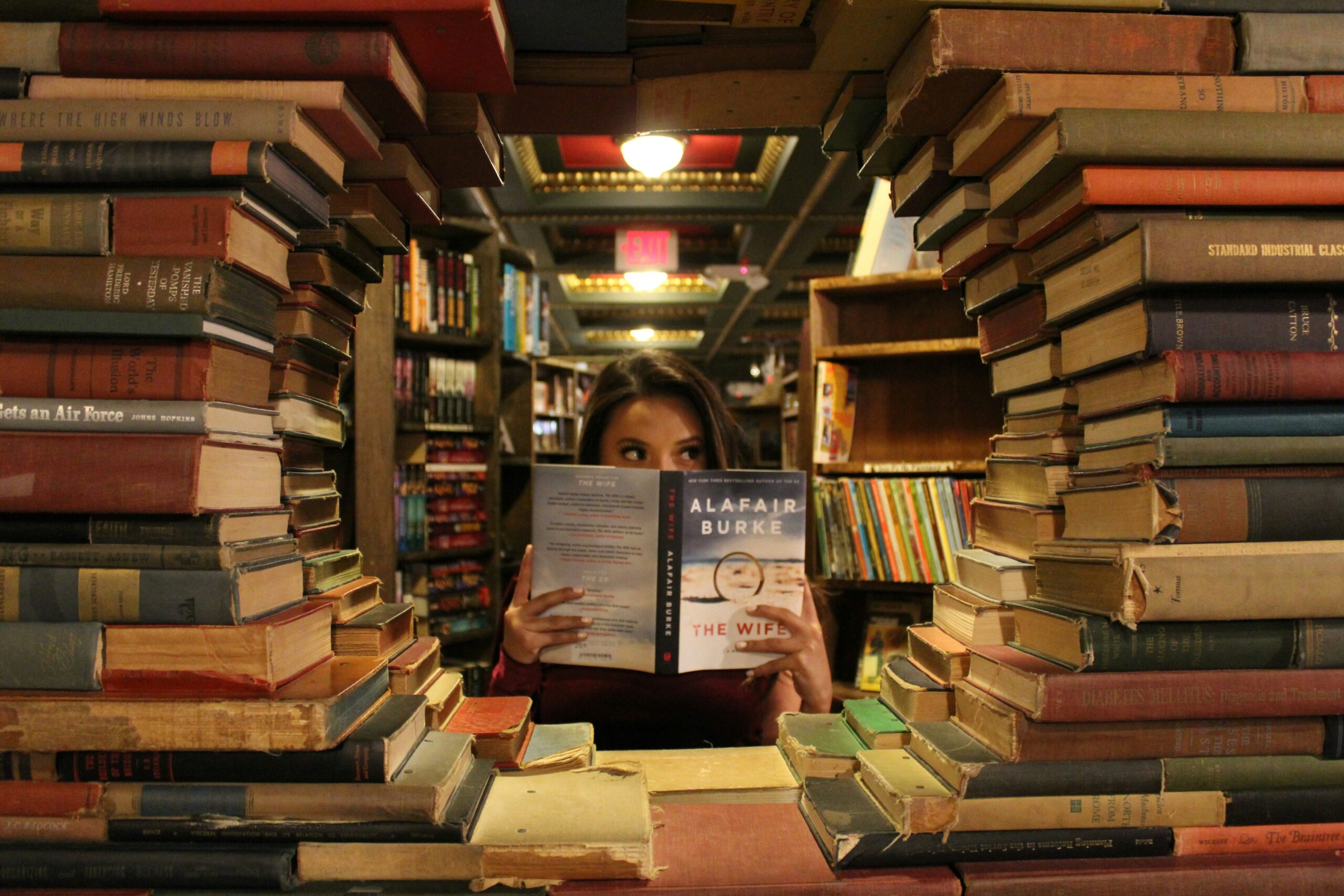A woman is reading IDP full form partially obscured by an open book she is holding, peeking over the top. She is framed by large, uneven stacks of books on both sides, creating a tunnel-like view into a warmly lit, slightly blurred background of a library or bookstore.