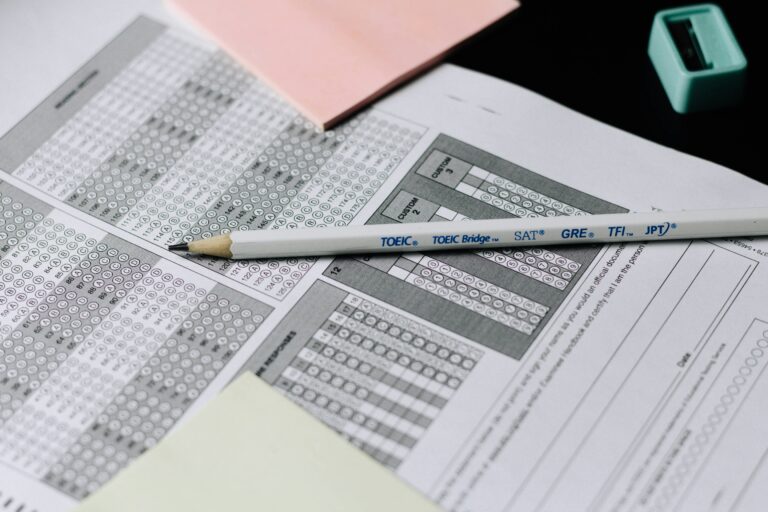 Close-up of a pencil resting on a standardized test answer sheet (bubble sheet) with sections for SAT, GRE, and TOEIC Bridge. A pink sticky note and a sharpener are visible in the background.